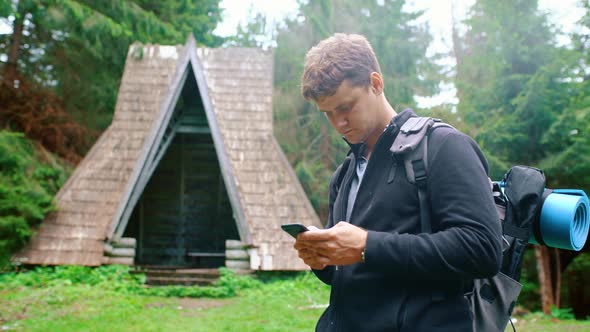 Young Man with a Backpack Stands in the Forest and Writes a Message on the Phone