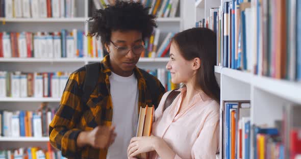 Young Multiethnic Couple Standing Against Bookshelf in College Library