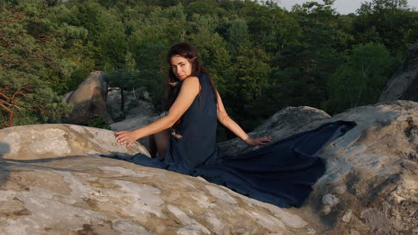 There is a Young Woman Sitting on Rocky Terrain Relaxing Outdoors While Wearing a Blue Silk Dress