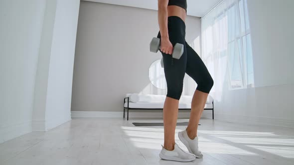 A Woman at Home on a Sunny Day in the Living Room Lunges Back with Dumbbells in Her Hands