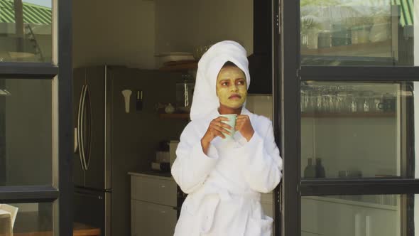 Happy african american woman with beauty mask on face, drinking coffee in kitchen