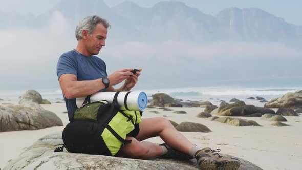 Senior hiker man with backpack using smartphone while sitting on rock hiking on the beach.