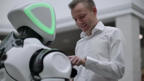 A Man in a Shirt Communicates with a White Robot Asking Questions and Pressing the Screen with His