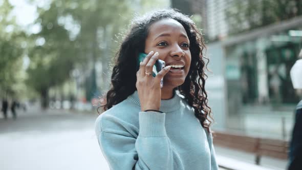 Cheerful brunette African woman wearing blue sweater talking by phone and walking