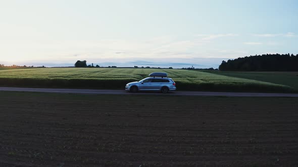 A Car with Roof Rack Drives Along a Road Through Green Meadows and Arable Fields