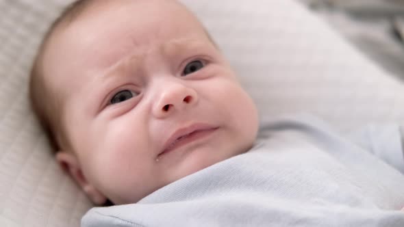 Dissatisfied Crying Upset Sad Newborn Baby Boy Lying in Nursery Cocoon with Blue Clothes on Bed in