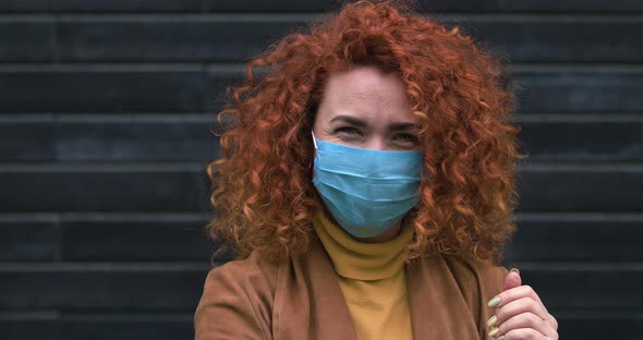 Beautiful Young Red-haired Caucasian Woman in Medical Mask Looking Into the Camera Touch Hair