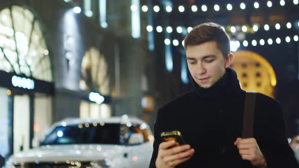 A Young Man Walks Through the Night City Uses a Smartphone