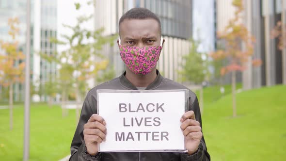 A Young Black Man in a Face Mask Shows a Black Lives Matter Sign To the Camera - Office Buildings