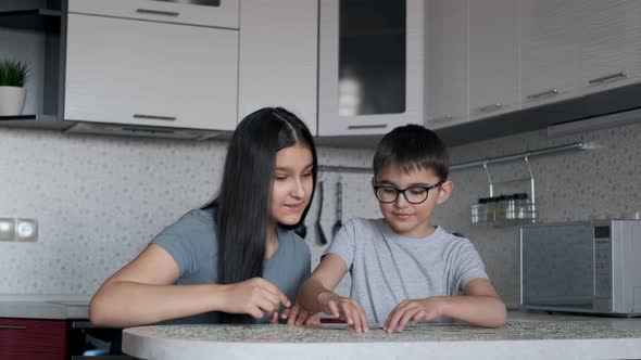 Boy and Girl Put Together a Puzzle While Sitting at a Table at Home in the Kitchen