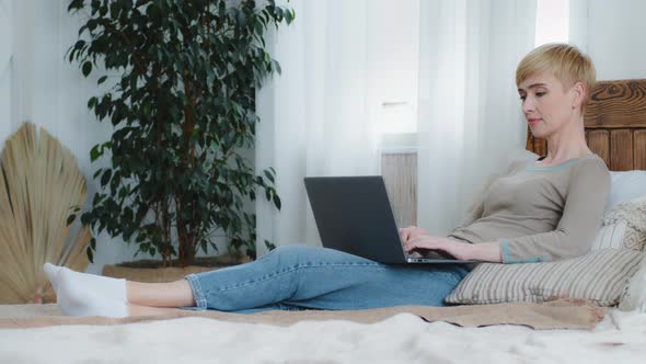 Smiling Relaxed Millennial Woman Lying on Comfortable Bed with Computer on Laps Typing Communicating