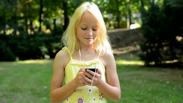 Little Cute Girl Listen Music in the Park on the Smartphone Through the Headphone