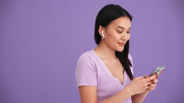 Positive Asian brunette woman in headphones typing by phone