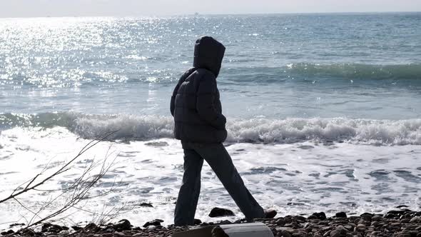 Unrecognizable Lonely Woman in Down Jacket and Jeans Walks Along Sea or Ocean Along Stone Beach