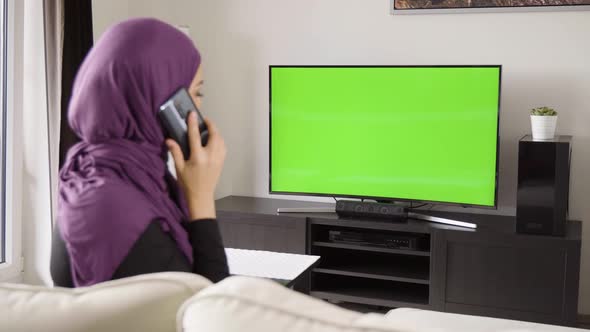 A Muslim Woman Talks on a Smartphone As She Watches TV with Green Screen in an Apartment