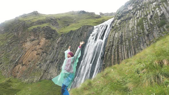 Lady Jumps Up and Waves Hands Standing Against Waterfall