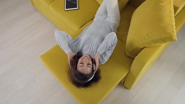 Gorgeous Cheerful Woman with Headphones Laying on the Cozy Sofa and Enjoying Music