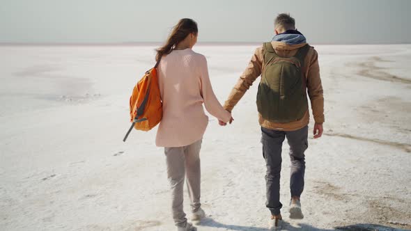 Couple Hikers with Backpacks Walking Holding Hands on Deserted Pink White Salty Coast of Sivash Pink