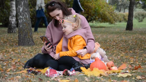 Mom with a Small Daughter Looking Photo on the Phone in the Autumn City Park