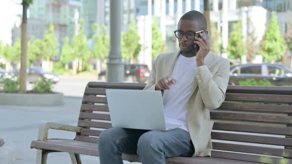 African Man Talking on Phone and Using Laptop While Sitting Outdoor on Bench