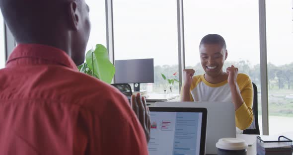 Happy african american creative businesswoman celebrating at desk in office with male colleague