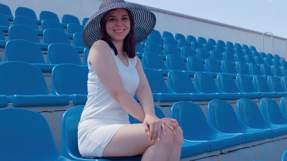 Young woman in white dress and elegant hat looks at camera, sitting on stadium bleachers alone.
