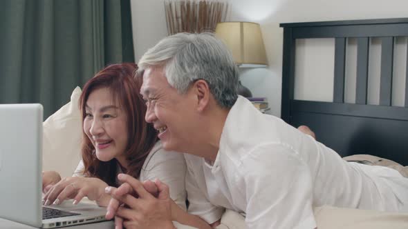 Asian senior couple using laptop after wake up, watching movie lying on bed in bedroom.