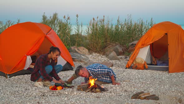 Young Loving Couple of Tourists Camping Sitting By the Fire Against Orange Tents on the Beach