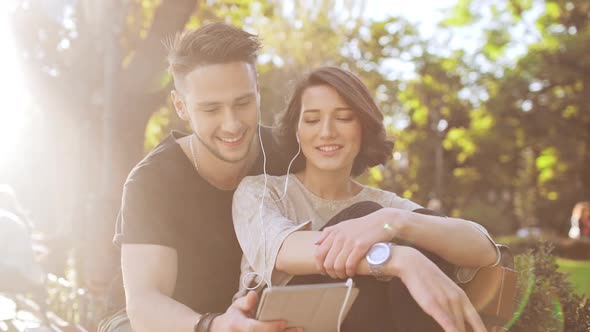 Young Beautiful Couple Smiling Speaking Looking at Tablet Sitting in City Park