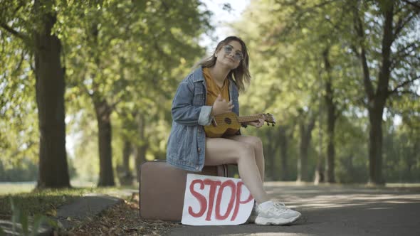 Wide Shot Positive Hippie Woman Sitting Suitcase Stop Sign Playing Ukulele Singing
