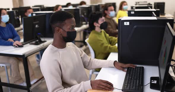 Young multiracial students using computers inside college class room while wearing safety masks