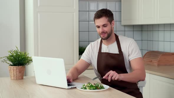 An Attractive Man Uses a Laptop While Sitting in the Kitchen and Eats a Vegan Salad