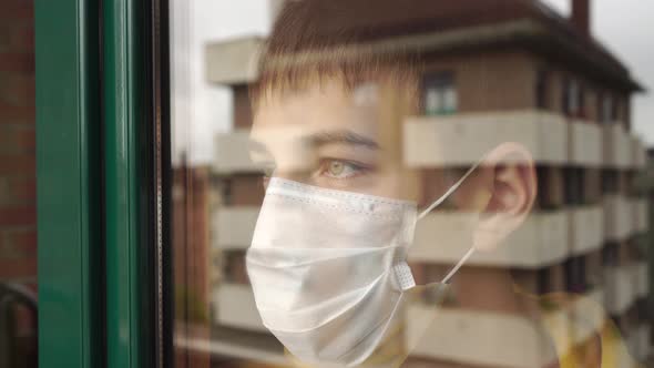 Teenager in a face mask in quarantine looks sadly out of the window and slaps his hand on the glass.