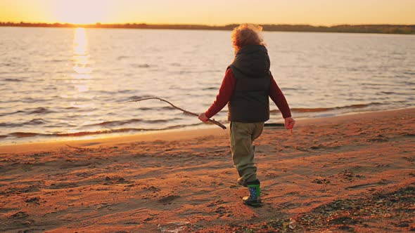 Cheerful Little Curly Boy is Running to Water of River in Sunset Time Walking on Sandy Coast in