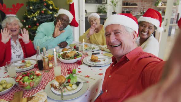 Happy diverse senior friends in santa hats taking selfie and waving at christmas dinner table