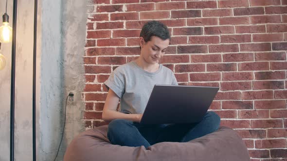 Smiling Woman Sits in a Room on Bag Chair and Working Behind a Laptop Looks Into the Camera