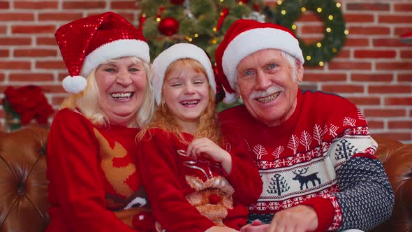 Senior Grandparents with Granddaughter in Santa Hats Laughing Out Loud at Home Near Christmas Tree