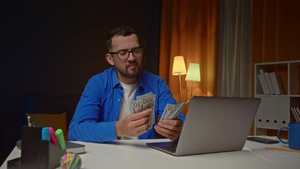 Young Man Counting Money Happily Sitting in Front of the Laptop Indoors in the Office on the