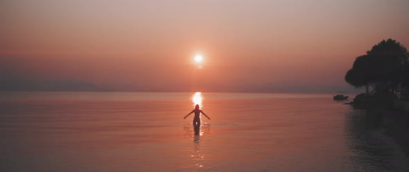 Silhouette of a young woman standing in a calm sea raising her arms in front of the sunrise