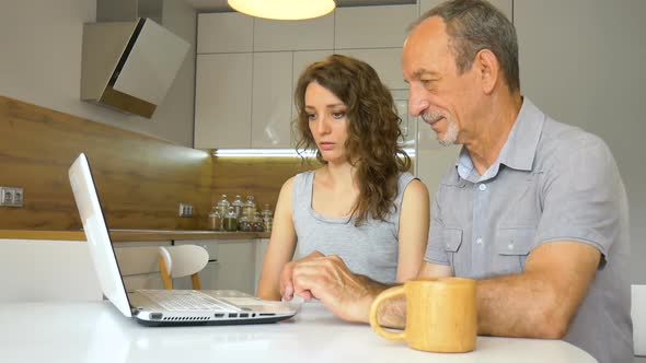 Attractive Young Daughter and Senior Father are Using Laptop Sitting in Modern Kitchen in Apartment