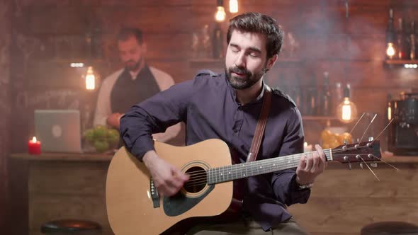 Hamdsome Bearded Man on a High Chair Plays and Sings on an Acoustic Guitar