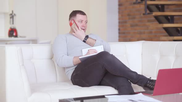 Wide Shot of Confident Adult Man Sitting on Couch Talking on the Phone and Writing in Workbook