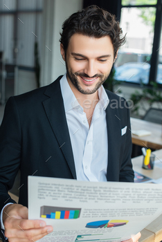 smiling businessman in white shirt and black blazer looking at analytics in office