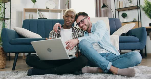 Mixed Race Male Partners Resting Together on the Floor at Home and Use Laptop