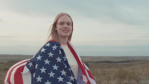 Beautiful Woman with Flag of America on Shoulders Smiling Looks at the Camera Standing the Field