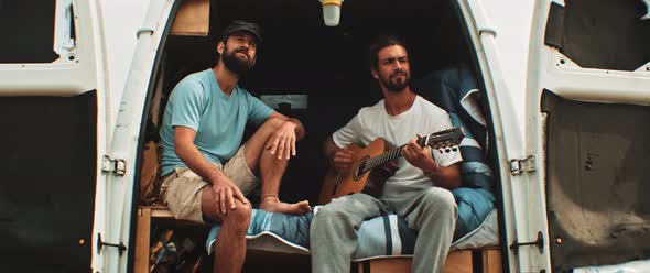 Two men sitting in back of a van singing and playing the guitar