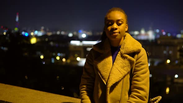 A Young Black Woman Nods at the Camera with a Smile in an Urban Area at Night  City Lights