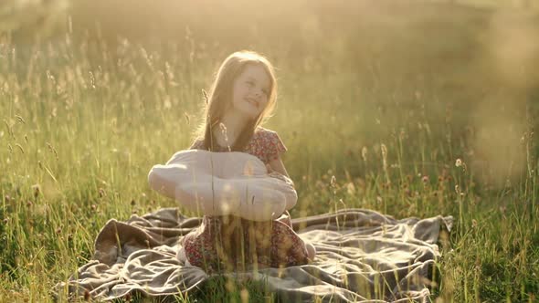 Little Cute Girl with Flowing Hair in a Summer Dress Sitting on the Grass and Playing with a Pink