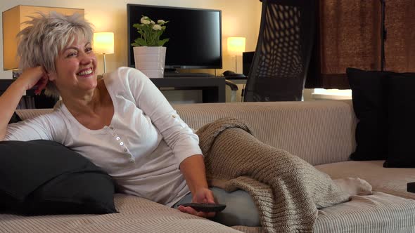 A Happy Middle-aged Woman Lies on a Couch in an Apartment Living Room, Watches TV