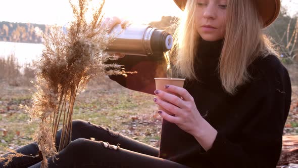 Caucasian Blonde Woman with Beige Hat in Black Sweater Drinks Hot Tea From Thermos in the